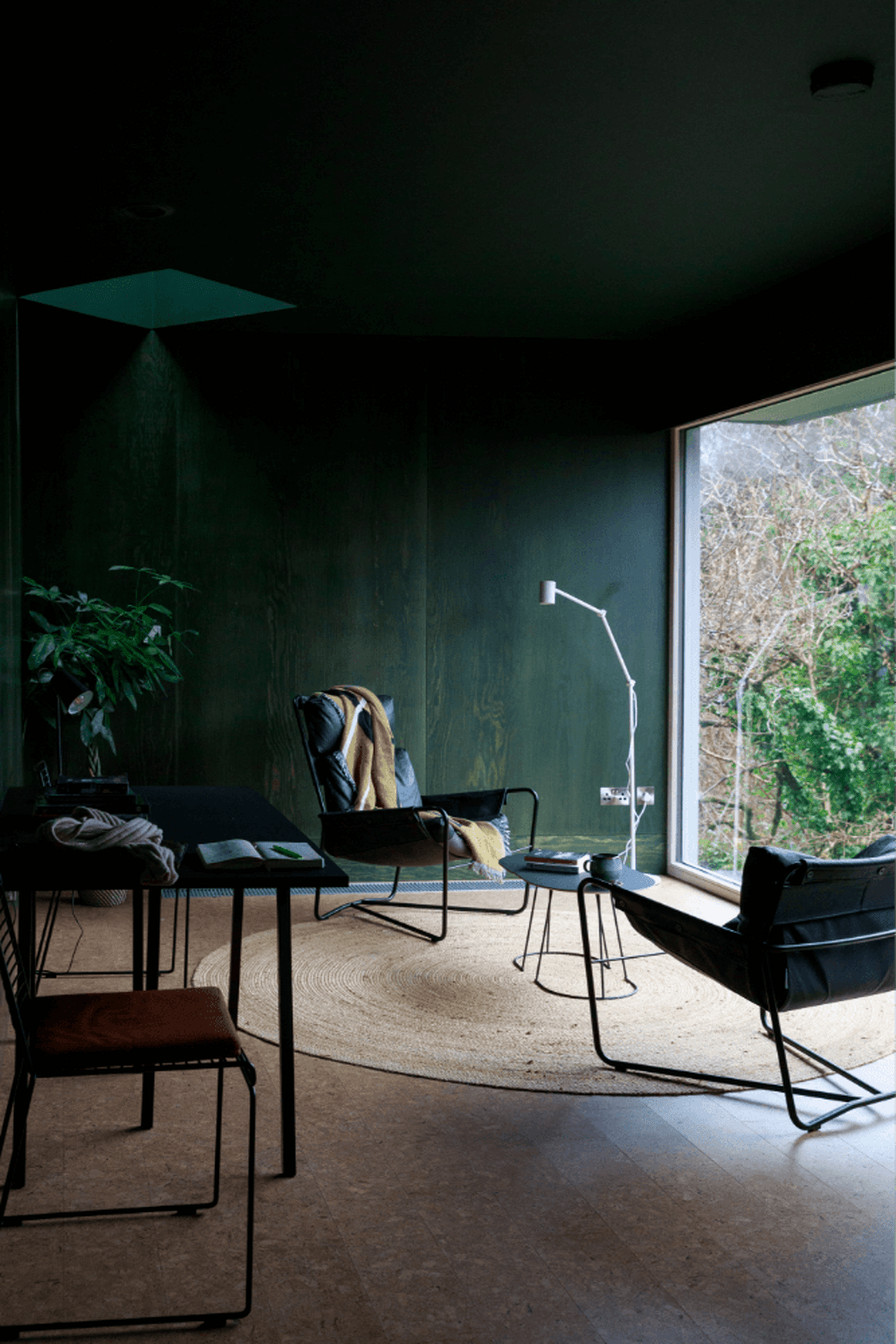 A forest green-tinted hotel room features wooden walls and leathery mid-century modern designs, with floor-to-ceiling windows letting nature step in.
