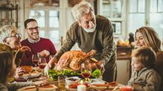 An older man sets a cooked turkey on the dinner table while his family members smile.