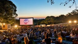 Lots of people sitting in front of a big screen that says Moonlight Cinema with sunset and trees in the background