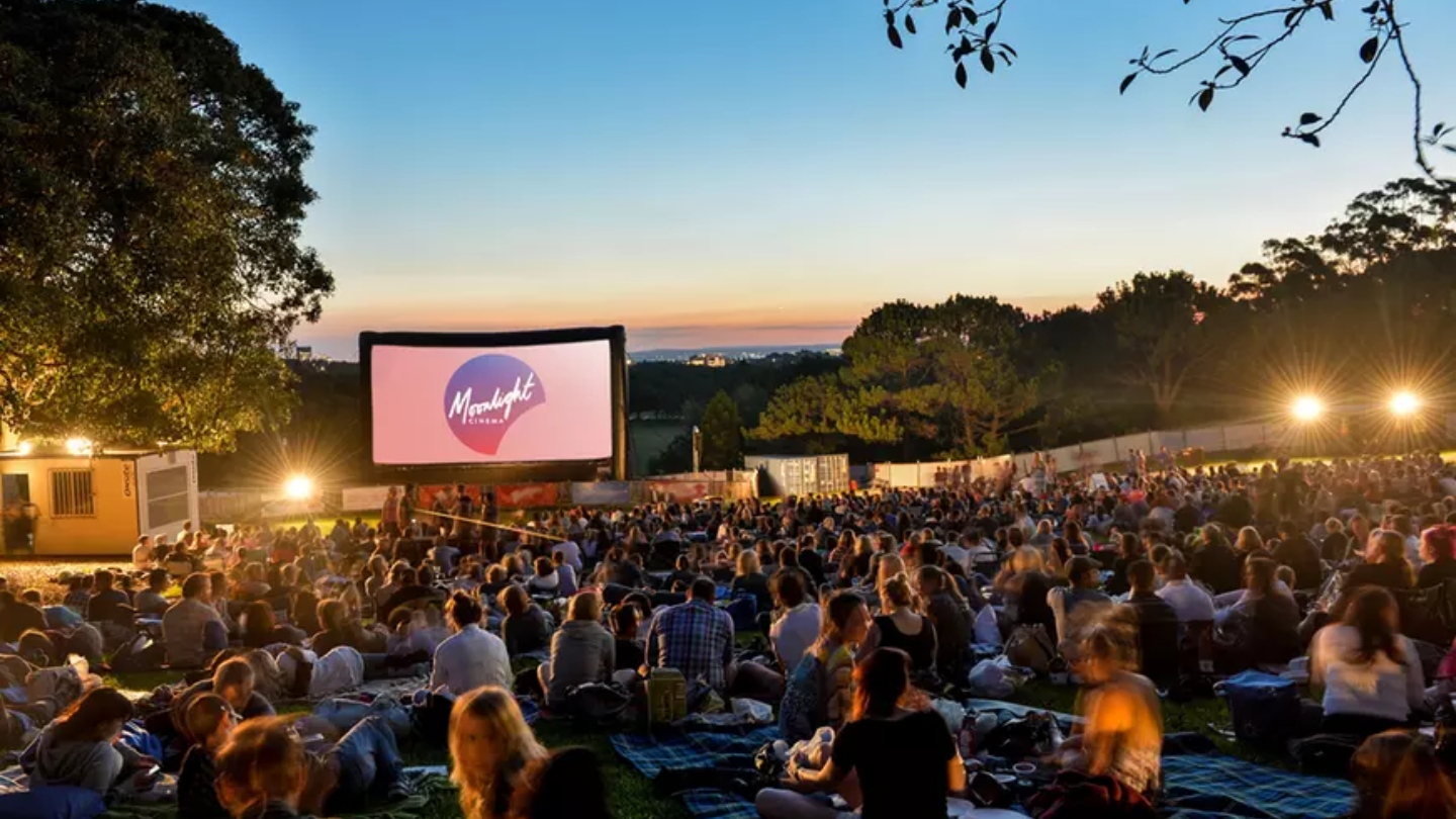Lots of people sitting in front of a big screen that says Moonlight Cinema with sunset and trees in the background