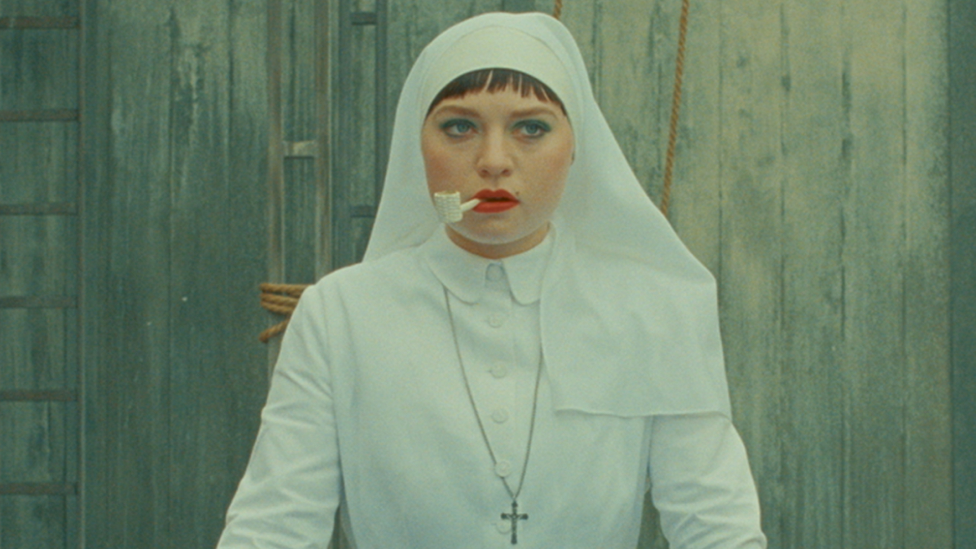 mia threapleton in a white nun habit smoking a pipe with bright red lipstick and green eyeshadow in the phoenician scheme