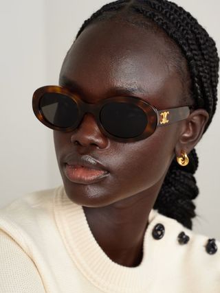 A woman wearing a beige jumper wears tortoise oval sunglasses with a side golden detail and a golden earring.