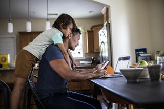 child hugging dad from behind while he looks at his phone and laptop