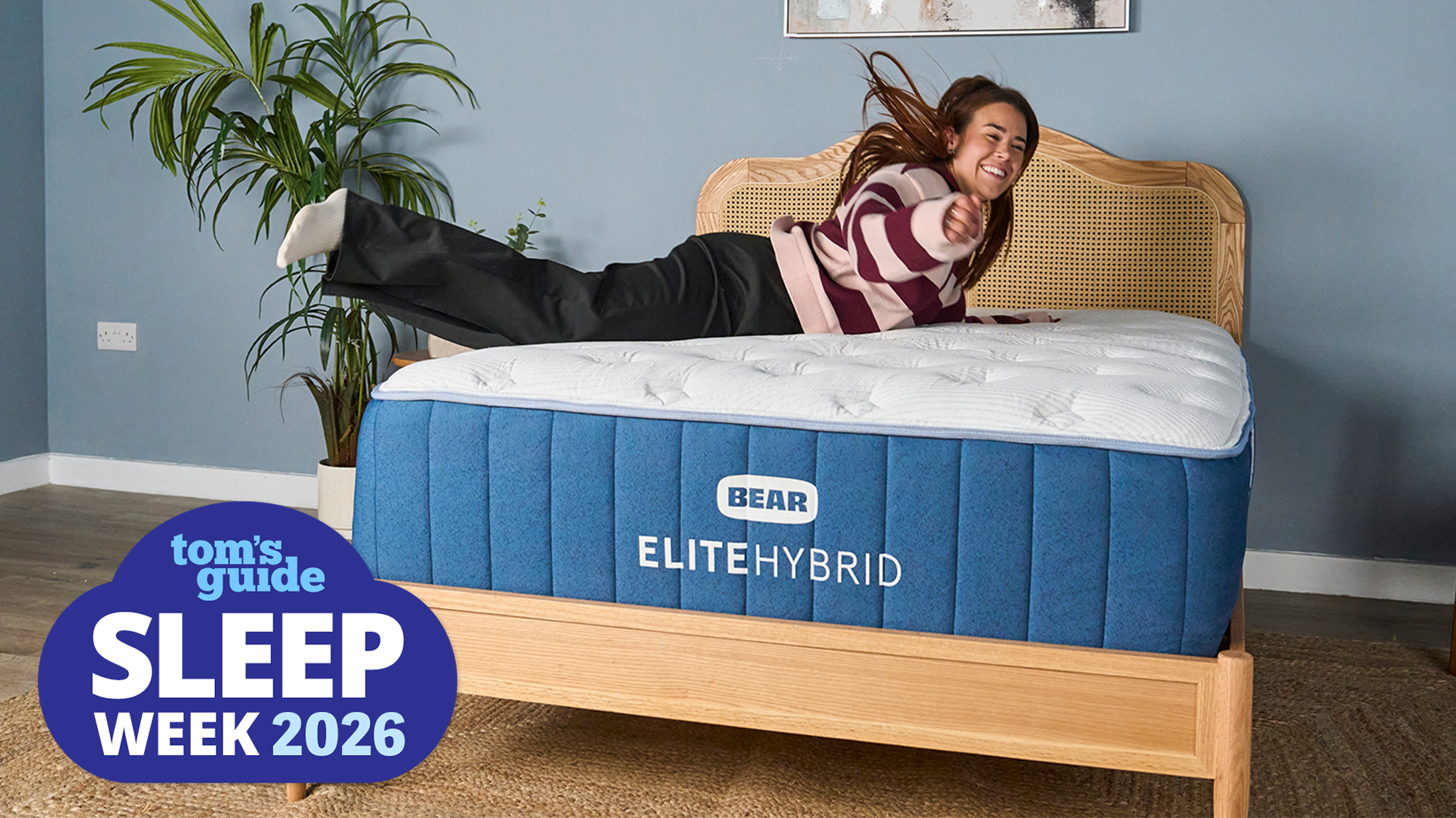 The Bear Elite Hybrid cooling mattress on a brown wicker bed frame, with Tom's Guide sleep tech writer Eve Davies jumping onto it after a great night's sleep. A blue Sleep Week 2026 logo, shaped to look like a cloud, sits in the bottom left hand corner.