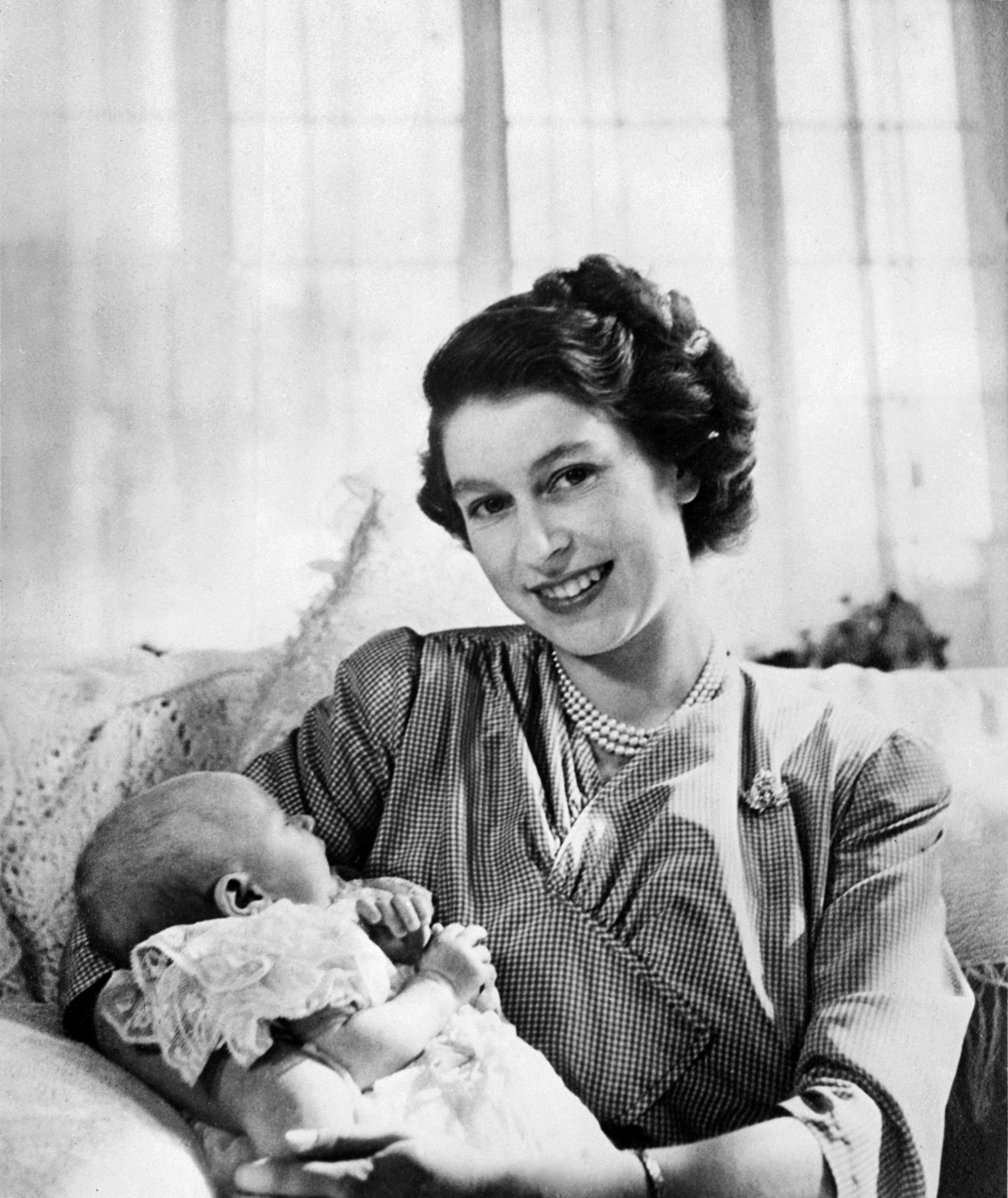 Queen Elizabeth holding baby Princess Anne in front of a lace curtain