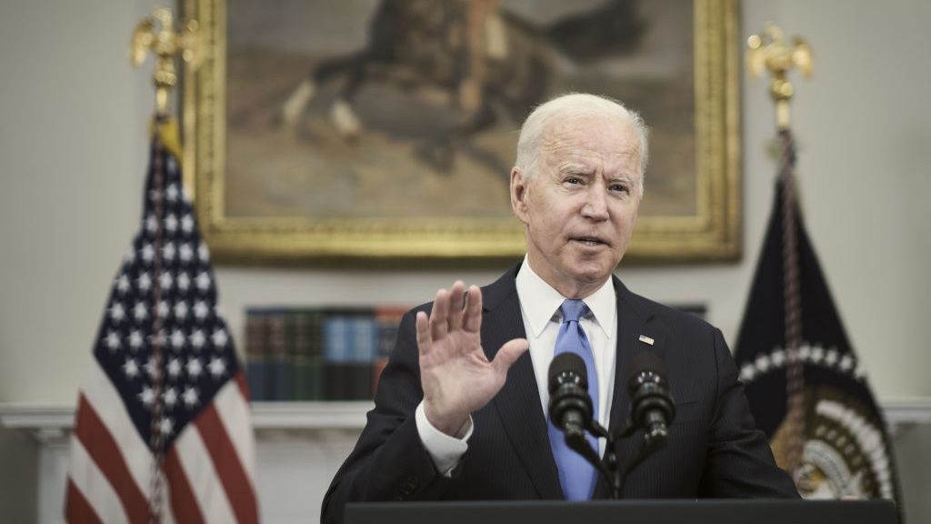 President Biden speaking into a microphone at a podium inside the Roosevelt Room of the White House, with his right hand raised.