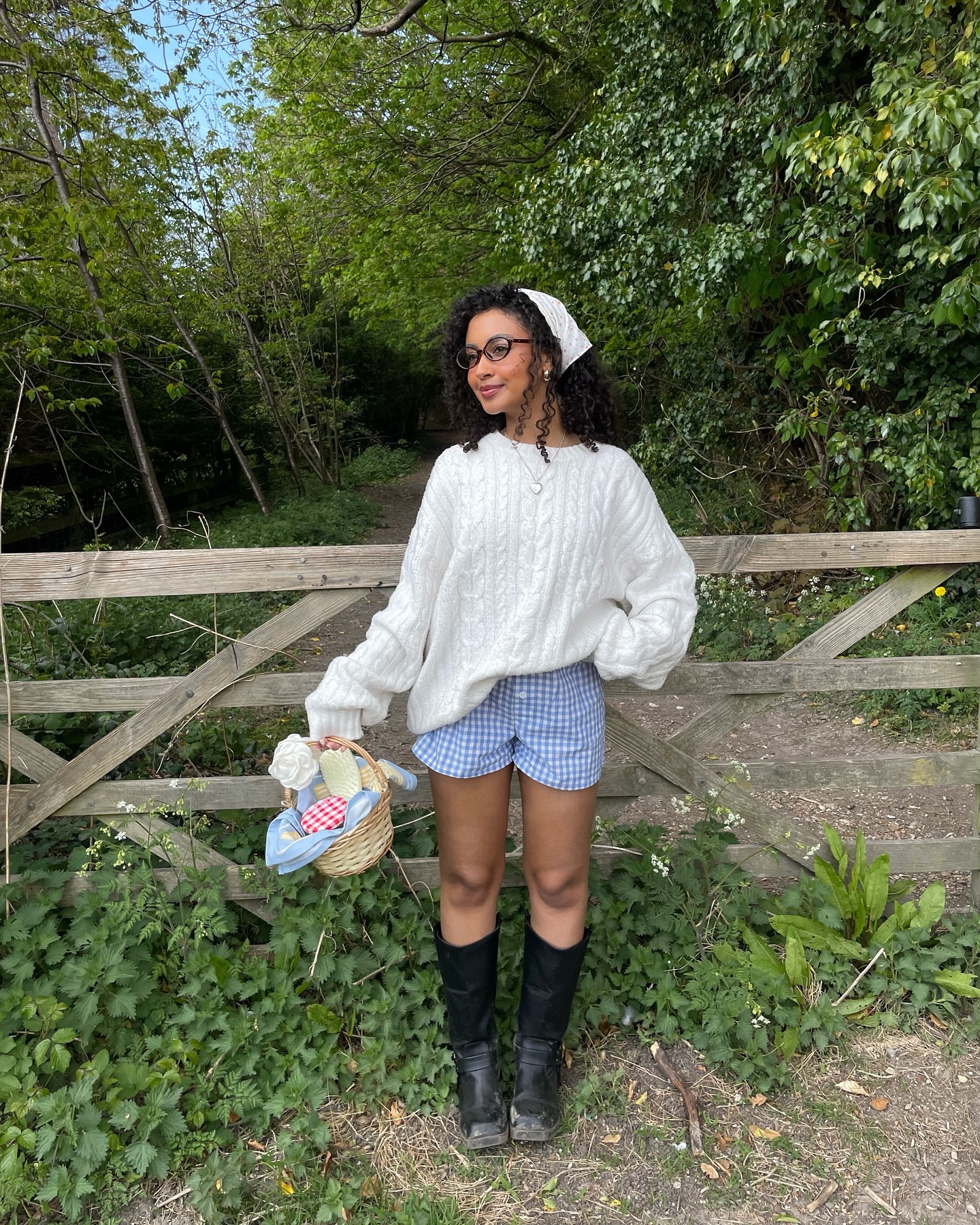 A woman wears a white cable sweater, blue boxer shorts, and knee high black boots.