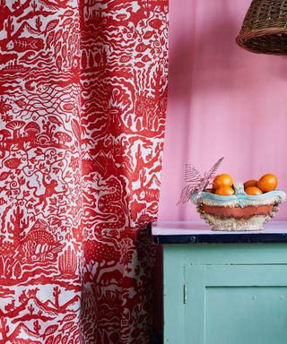 A close up of a teal blue console table with a blue bowl with oranges in it, a pink wall behind it, and a red and white patterned curtain panel to the left