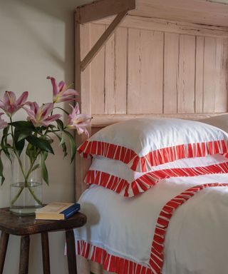 A pink bed frame with white bedding with red trim, next to a vase of lilies on a nightstand