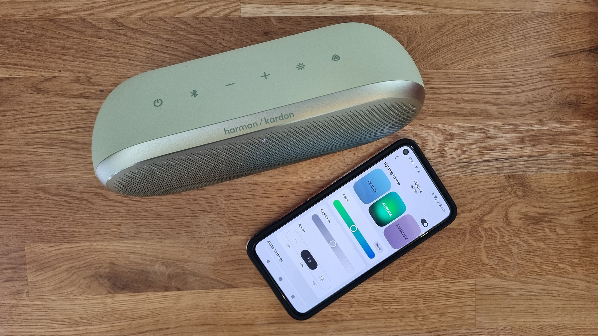 Harman Kardon Luna 2 wireless speaker on wooden table next to smartphone with app on screen