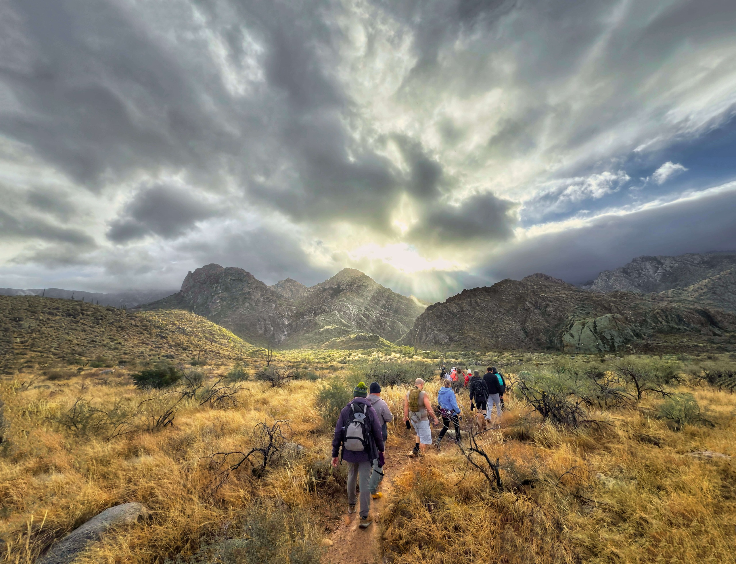 Hikers walking towards moody clouds with sunlight piercing through above a rugged landscape.