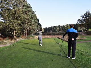 Sam De'Ath hitting a driver off the tee, watched by Nick Bonfield