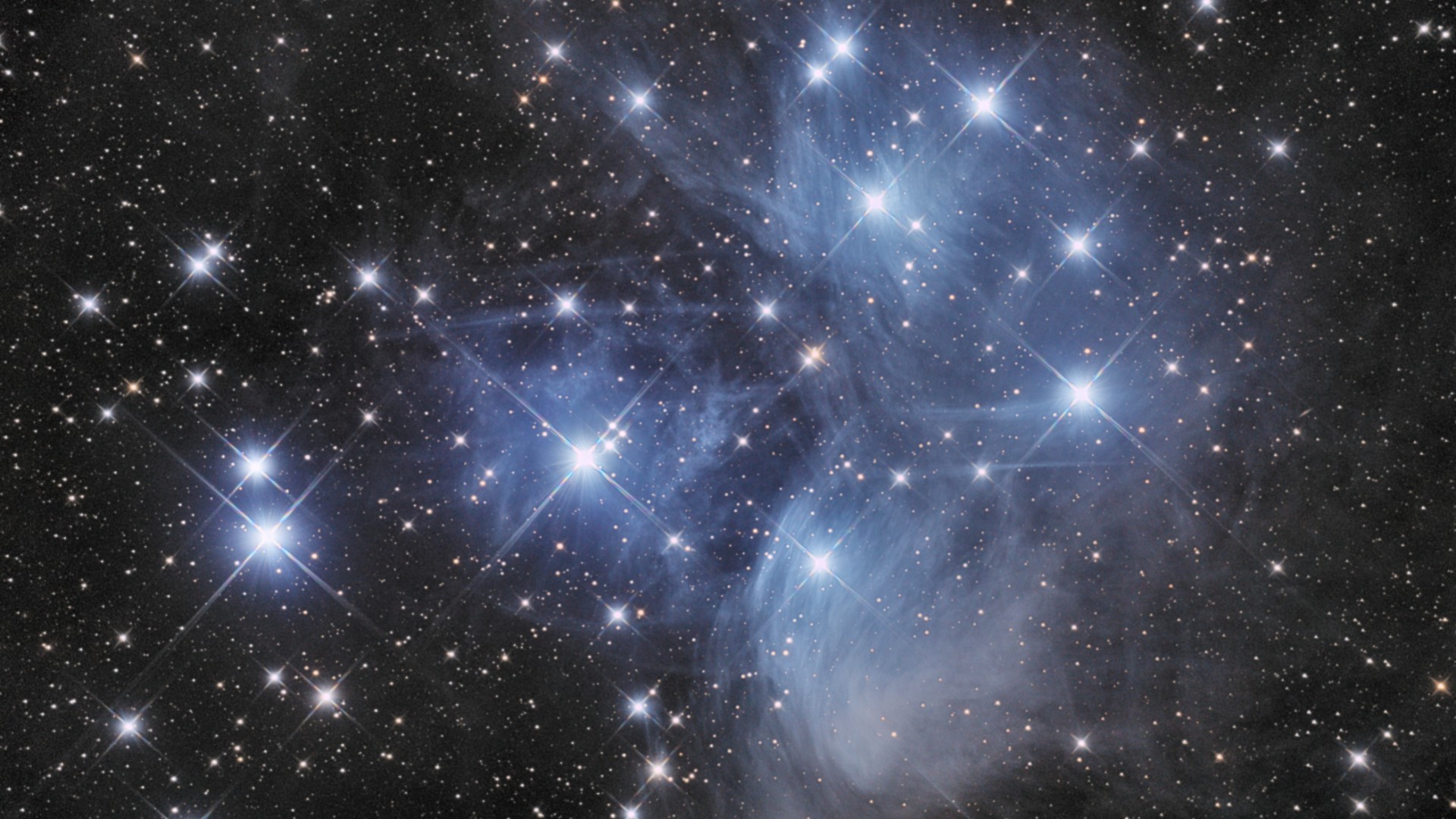 The Pleiades star cluster featuring bright blue stars surrounded by wispy, glowing interstellar nebulosity against a field of deep space.