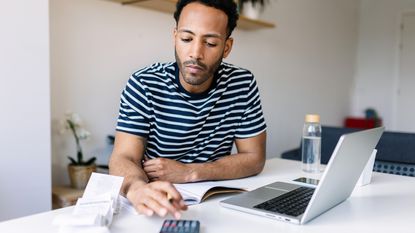 A man uses a calculator while sitting at a desk with his laptop open in front of him.