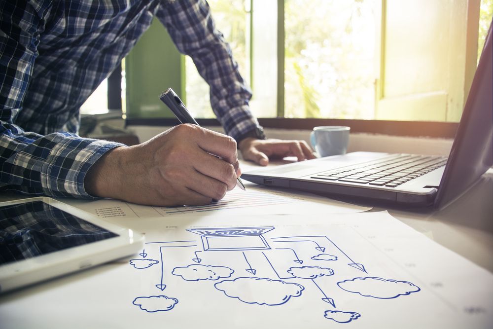 A person planning out a graph on a piece of paper showing a laptop surrounded by clouds