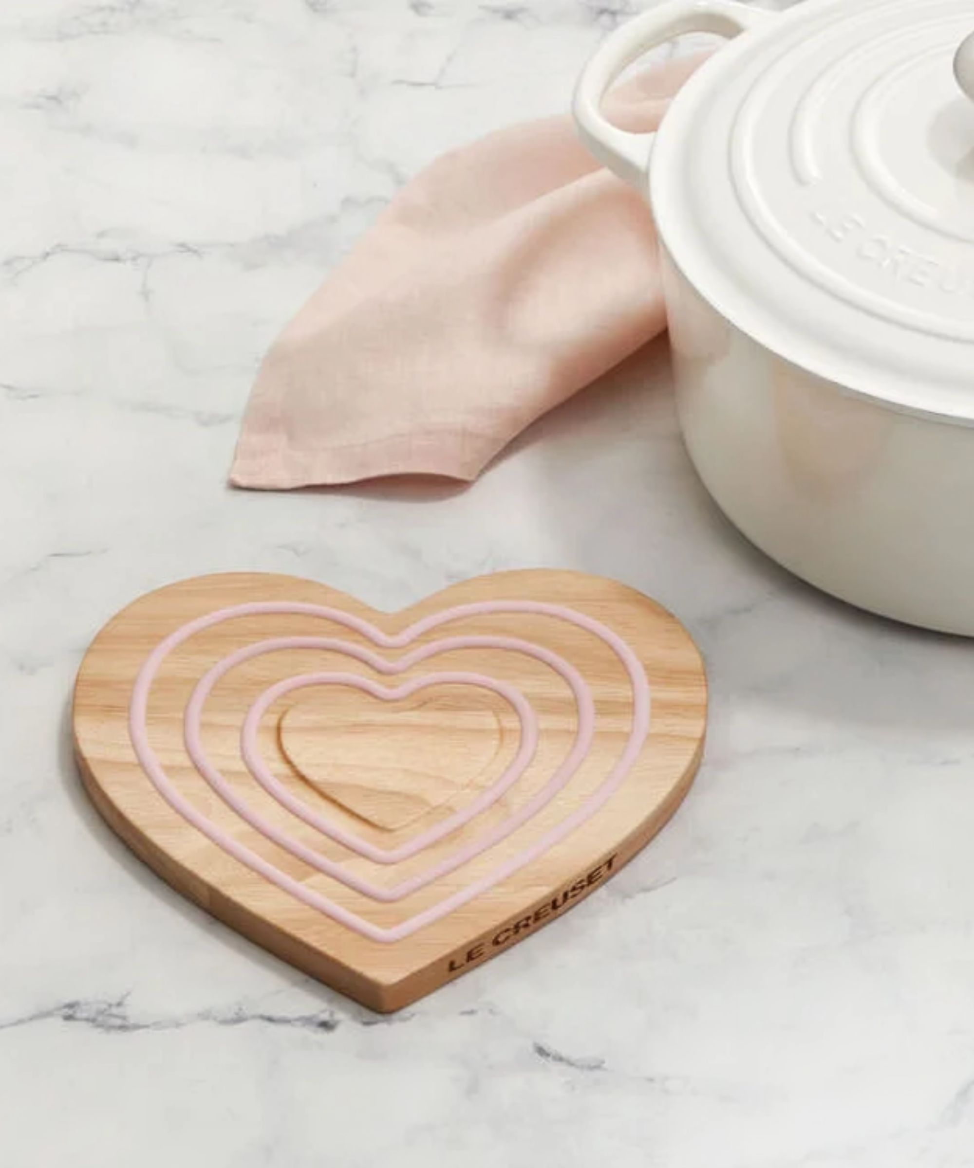 A wooden heart-shaped trivet for underneath hot pans, with three pink heart-shaped shapes set into it. On a white marble kitchen counter. The edge of a white ceramic Dutch oven beside it.