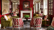 Bold living room featuring deep red walls, two olive green velvet sofas facing a fireplace, and two armchairs with red and white patterned fabric on a leopard print rug