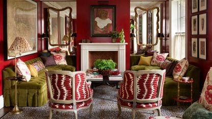 Bold living room featuring deep red walls, two olive green velvet sofas facing a fireplace, and two armchairs with red and white patterned fabric on a leopard print rug