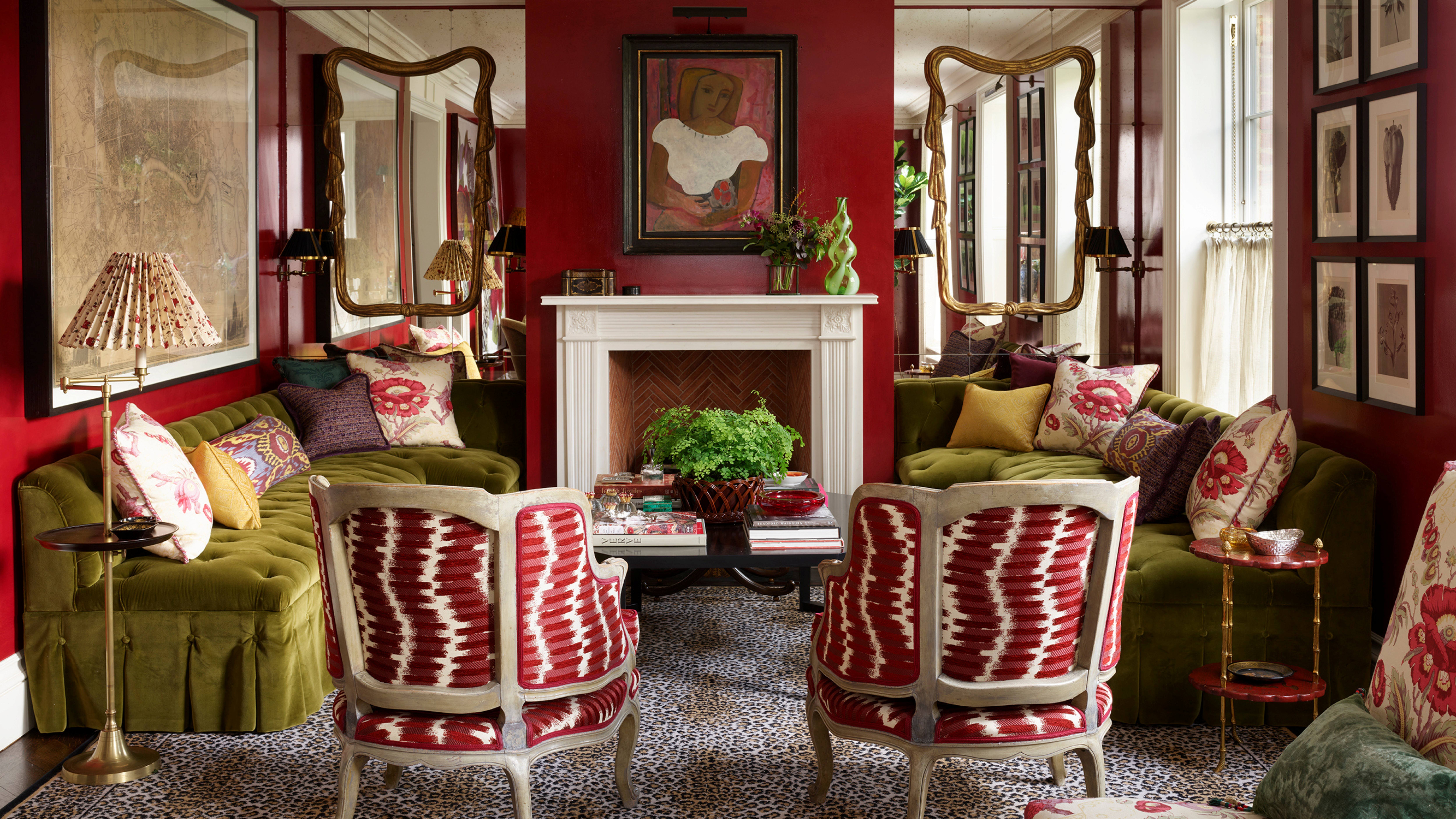 Bold living room featuring deep red walls, two olive green velvet sofas facing a fireplace, and two armchairs with red and white patterned fabric on a leopard print rug