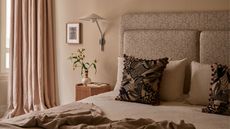 neutral bedroom with cream walls, beige curtains, a patterned upholstered headboard, wall sconce, timber side table with vase of flowers and picture frame, and bed made with natural linen and patterned cushions and throw 