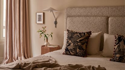 neutral bedroom with cream walls, beige curtains, a patterned upholstered headboard, wall sconce, timber side table with vase of flowers and picture frame, and bed made with natural linen and patterned cushions and throw 