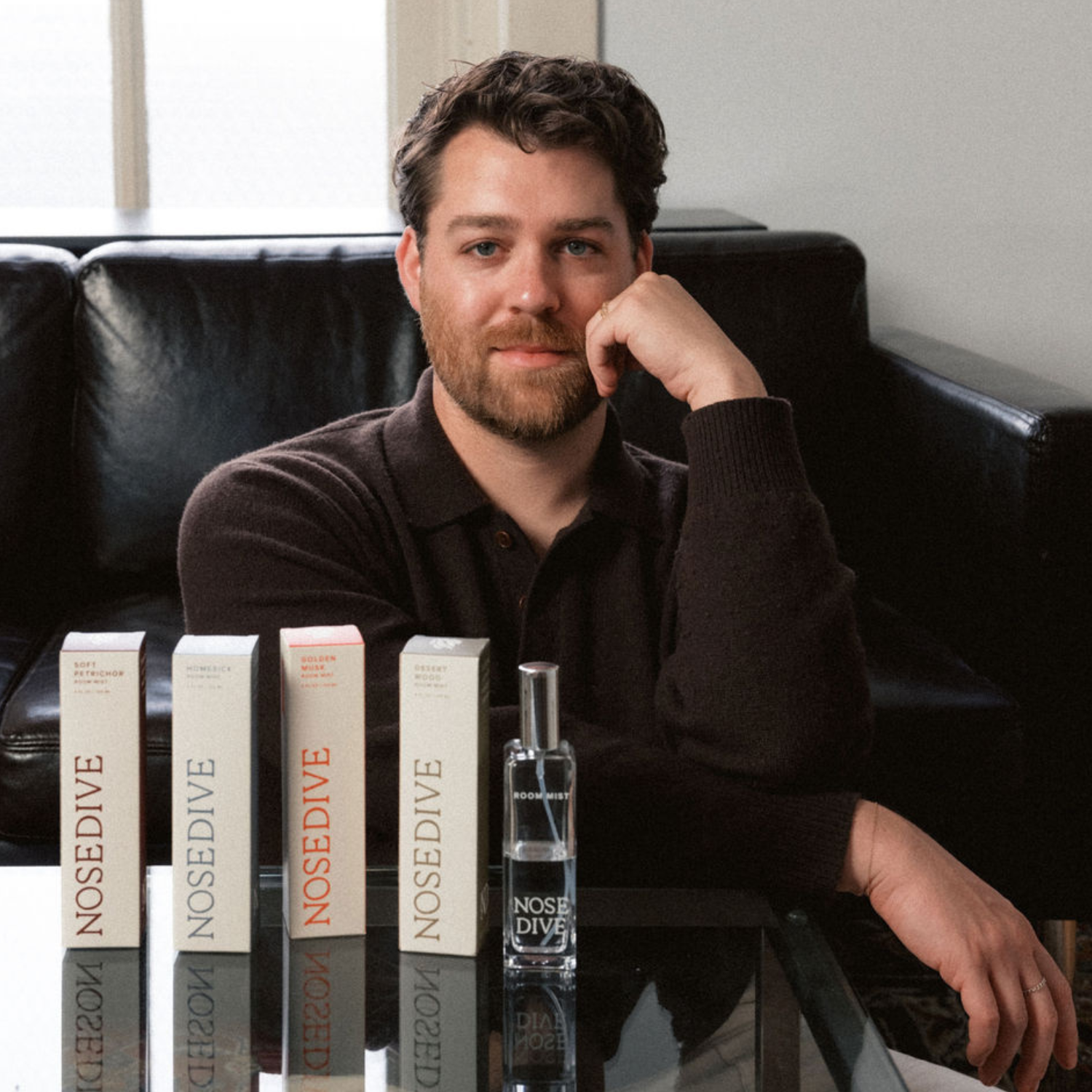 A headshot of a man with short brown hair in a brown sweater shirt sitting against a black leather sofa by a glass coffee table with glass room spray