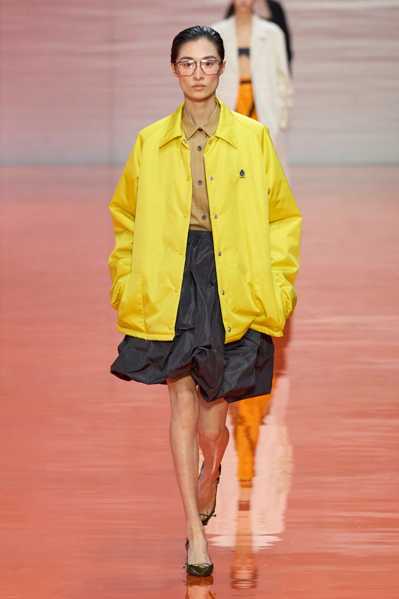 A Prada model wearing a citrus yellow collared jacket layered over a brown polo shirt, a black balloon skirt, black pointed pumps, and wire frame eyeglasses.