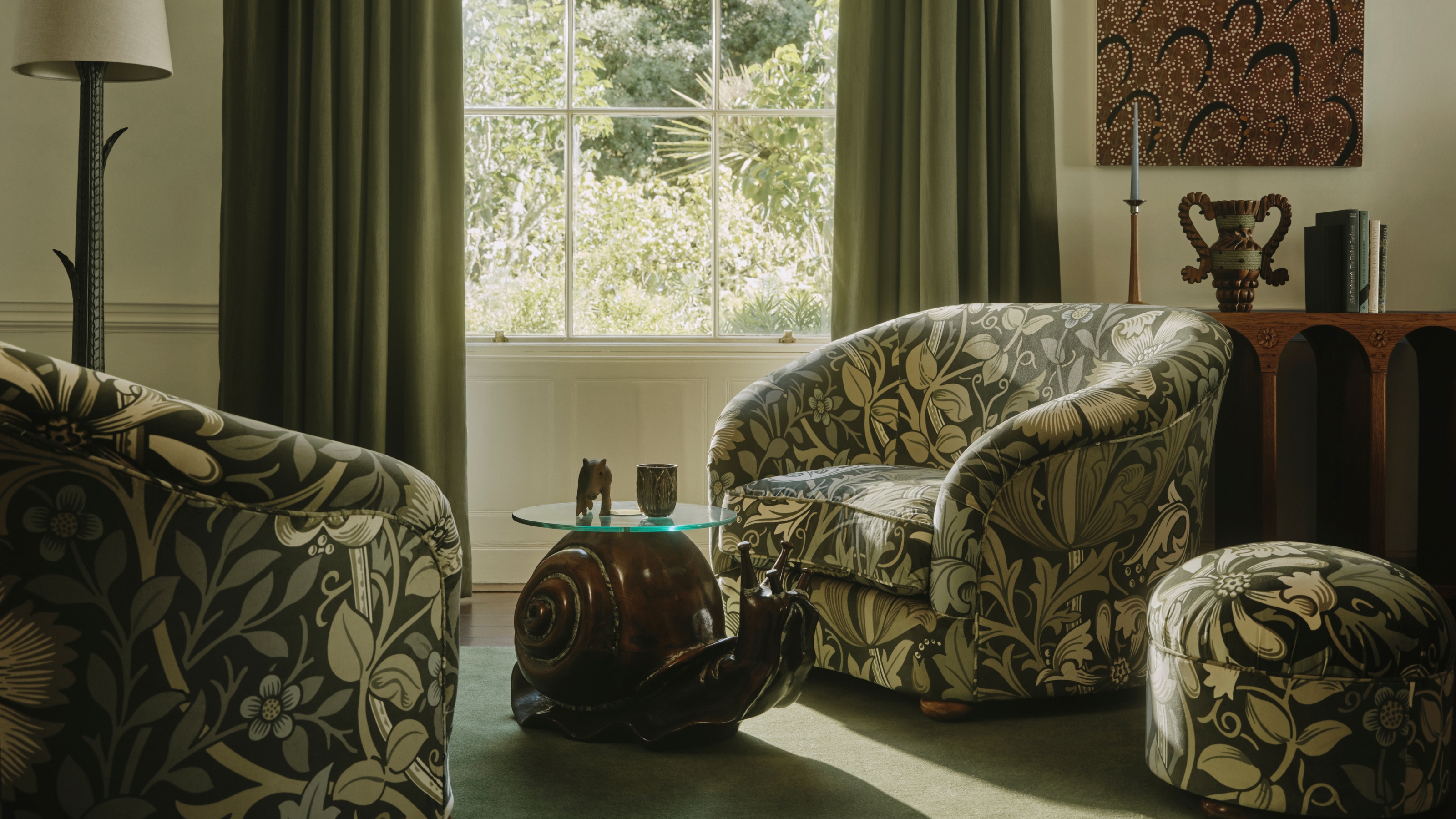 Image of a living room with a green color scheme. There are two matching accent chairs with a green floral pattern and an ottoman in the same pattern. In the middle of the chairs is a coffee table shaped like a snail.