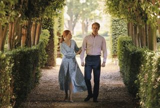 Princess Beatrice (Ellie Bamber) walks down a tree-lined path with her brother Prince Henry (Nicholas Galitzine). She has one hand on his arm consolingly, and his expression is burdened with troubles