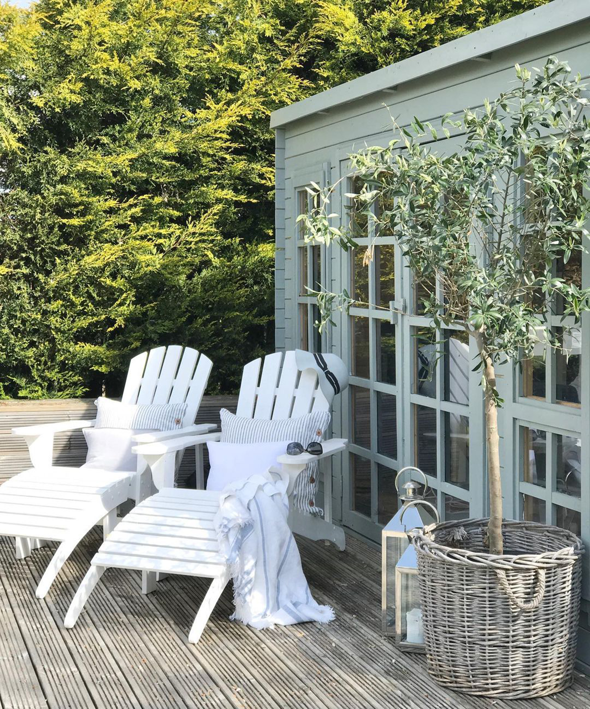 Two white Adirondack deck chairs on decking in backyard with rattan potted tree and lantern lighting