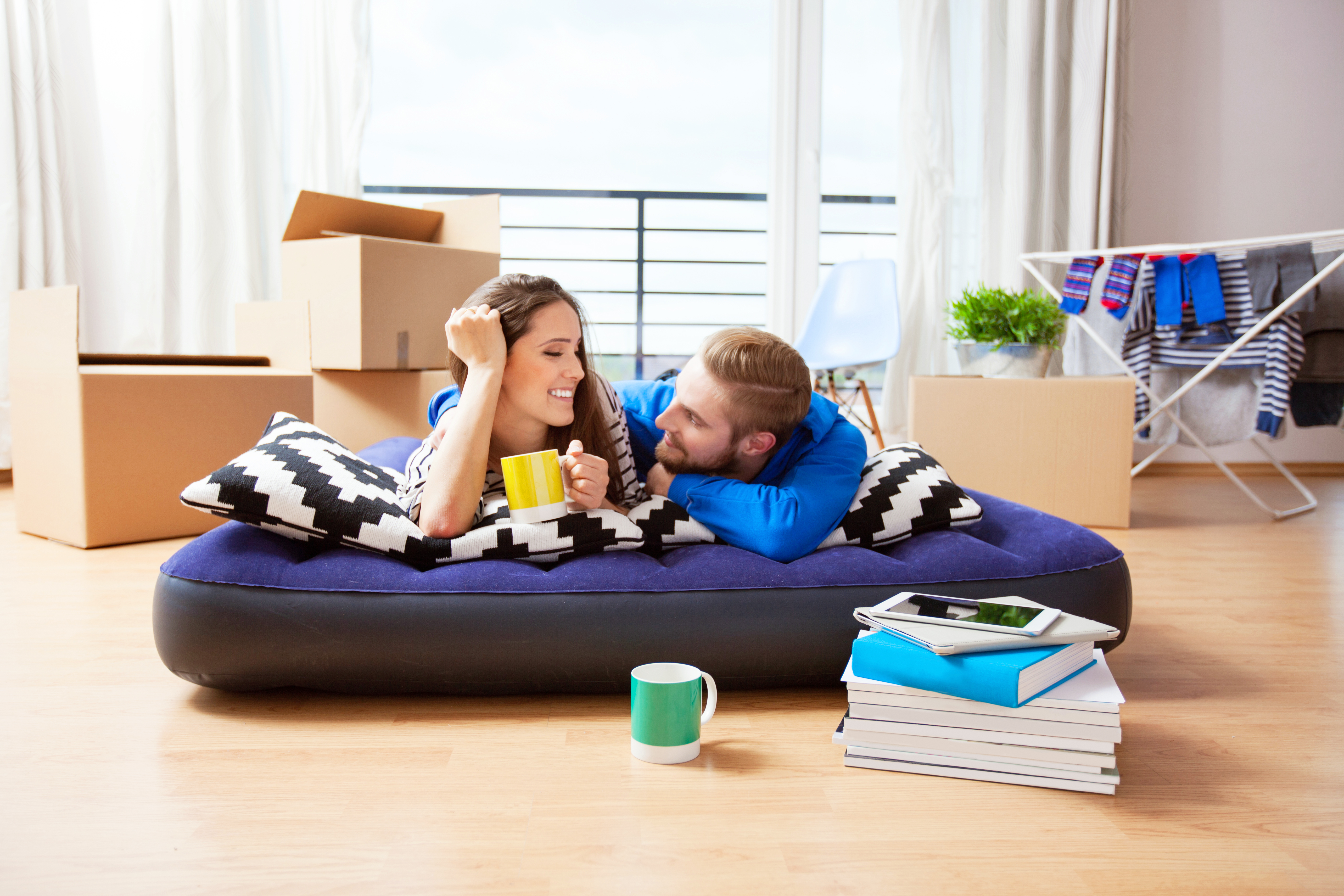 A young couple are relaxing on an inflatable mattress on the hard floor of a room with cardboard boxes on moving day.