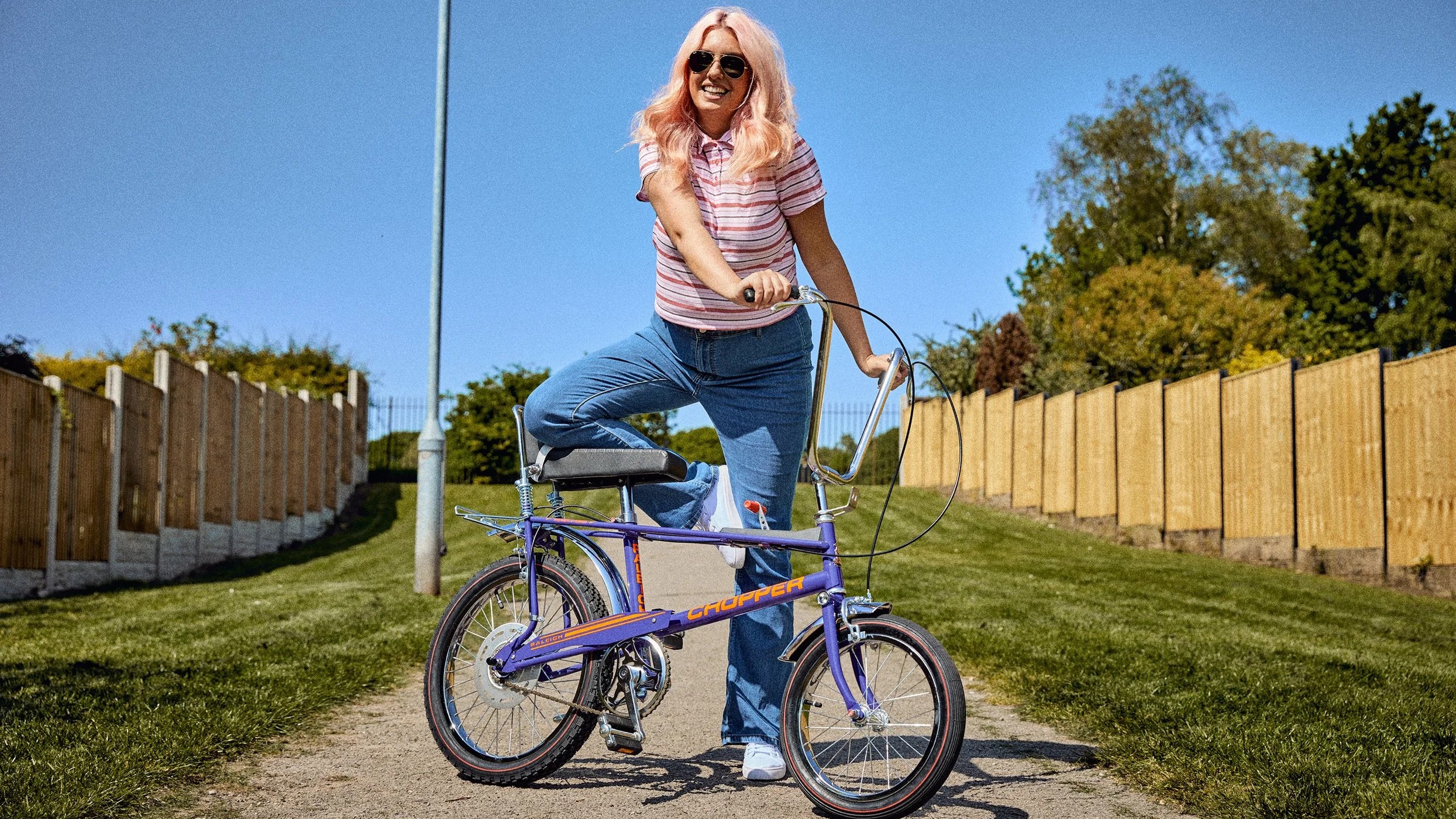 A woman with long blonde hair and sunglasses poses outdoors, leaning on a purple Raleigh Chopper bicycle.