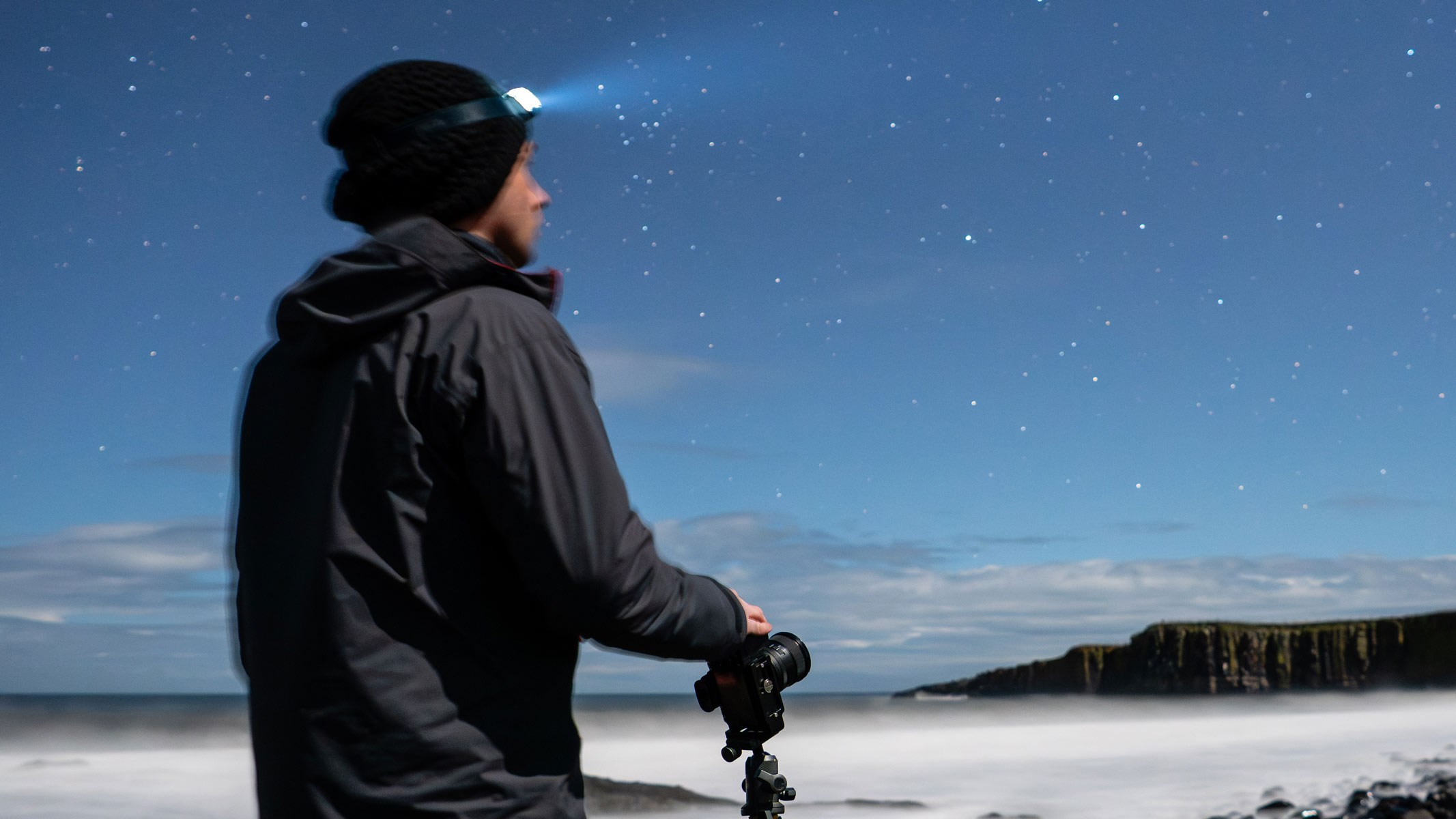 A man with a headtorch on and a camera in his hand with a backdrop of stars.