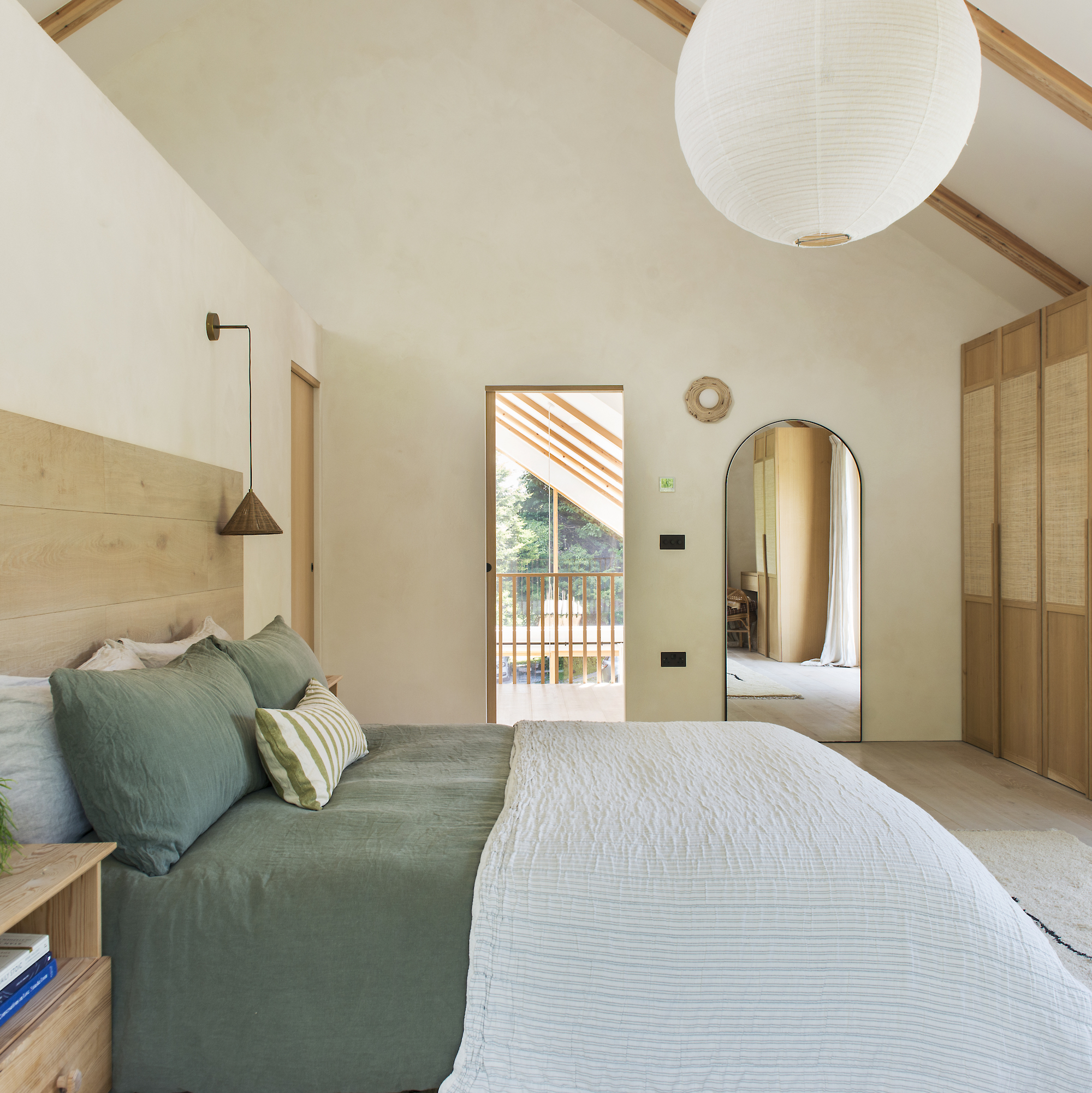 bedroom in contemporary house with vaulted ceiling and cement walls, with doorway to balcony. wooden bed has green linen bedding.