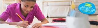 A pre-ten boy seated at a desk smiles as he writes on paper