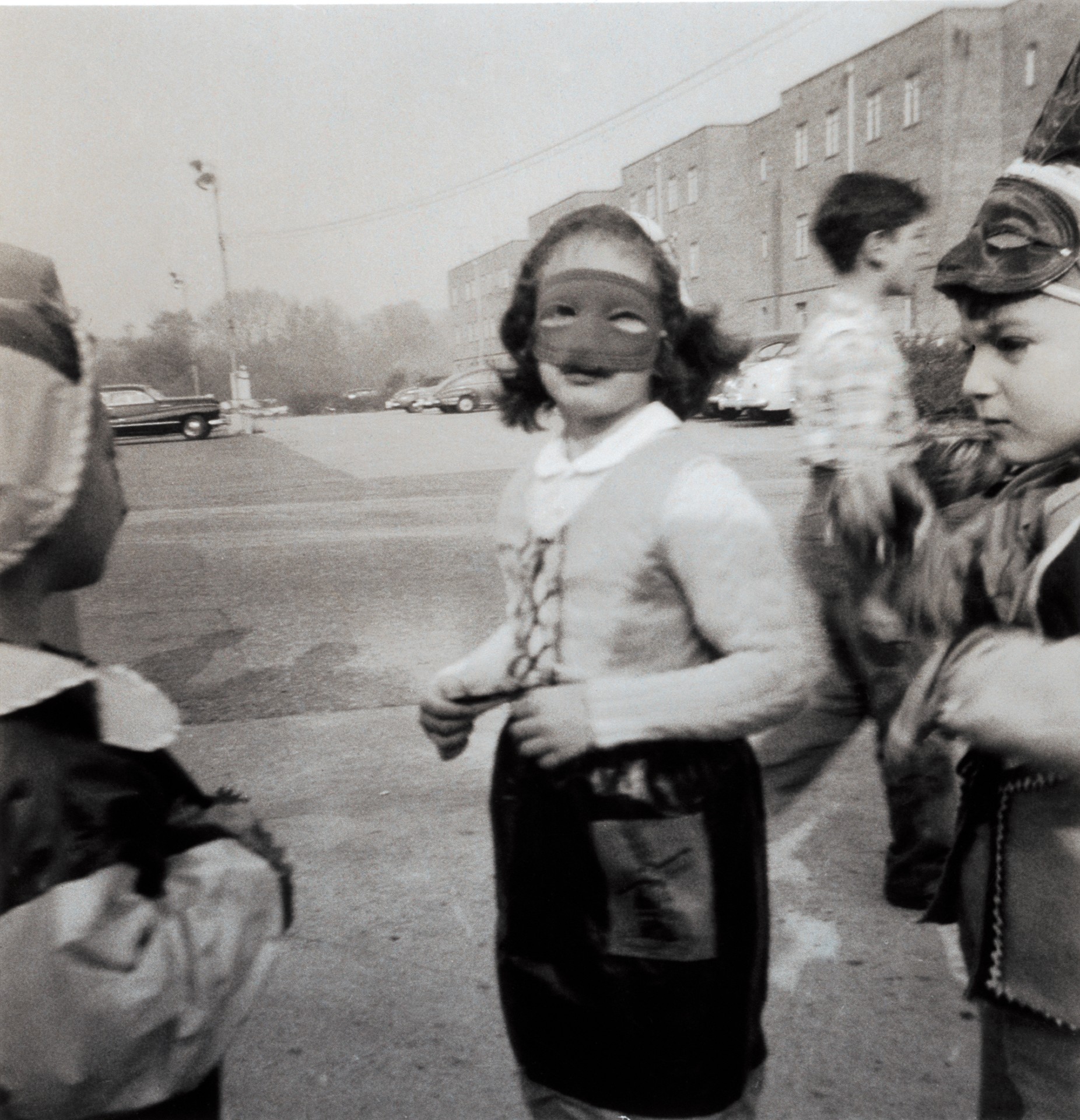 A black and white photograph of children in a parking lot wearing costumes and masks, including a girl in the center with a dark eye mask.