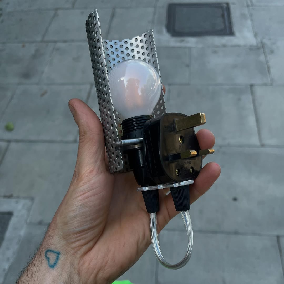 A hand with a heart tattoo holding a lamp. The lamp is made of a curved, perforated metal sheet shading a lightbulb, connected to a UK plug