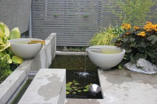 Solus Decor Water Bowl Scupper garden water feature in a paved garden with a pond and a grey fence