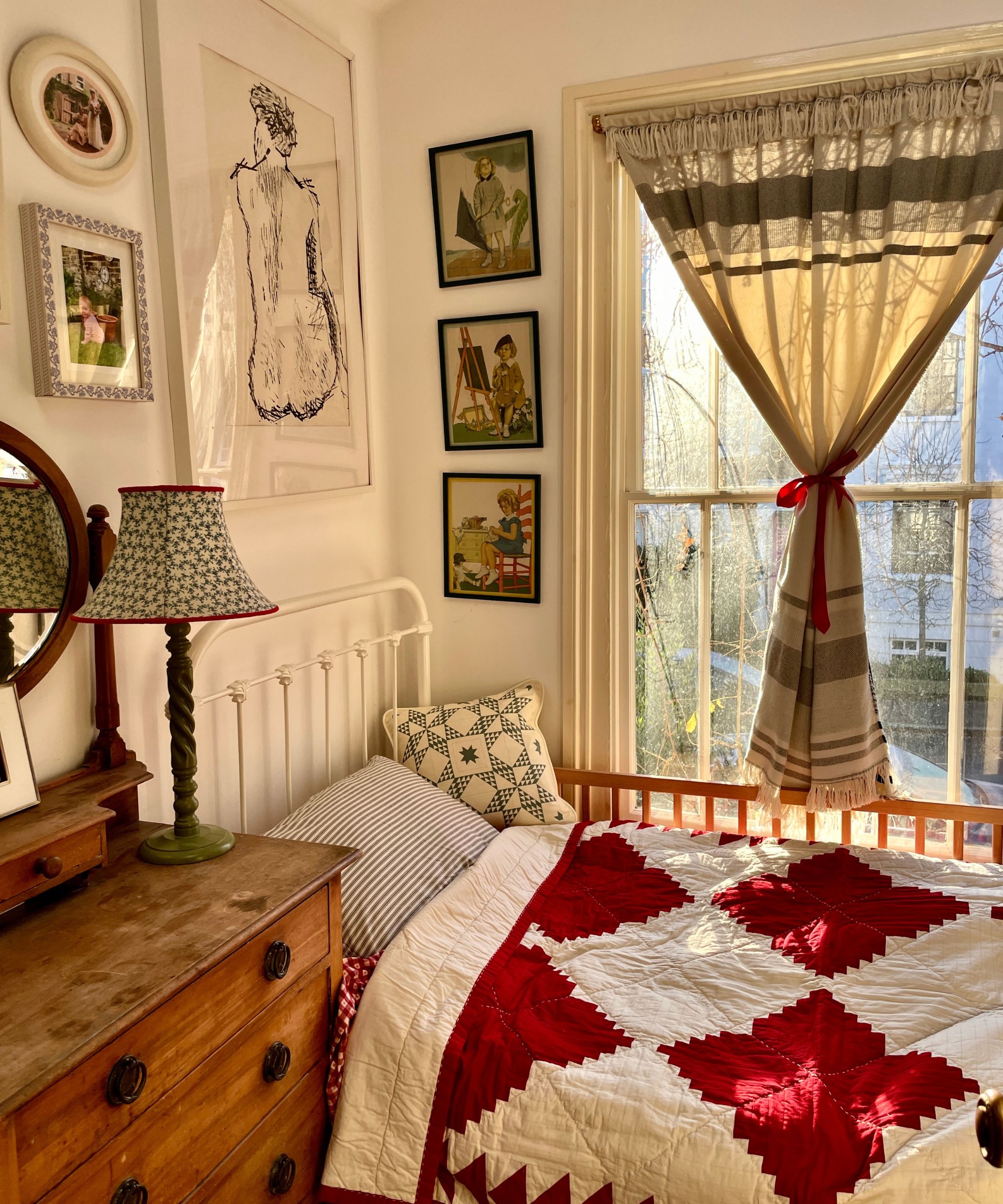 Sunny, eclectic bedroom featuring a white metal bed, a red and white quilt, a worn wooden dresser, and framed art hanging on the white wall