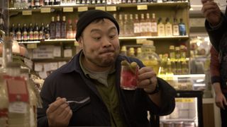 David Chang looks at a a glass mug he is holding with a red food in it, while holding a fork in his other hand and standing in a deli, in 'Ugly Delicious.'