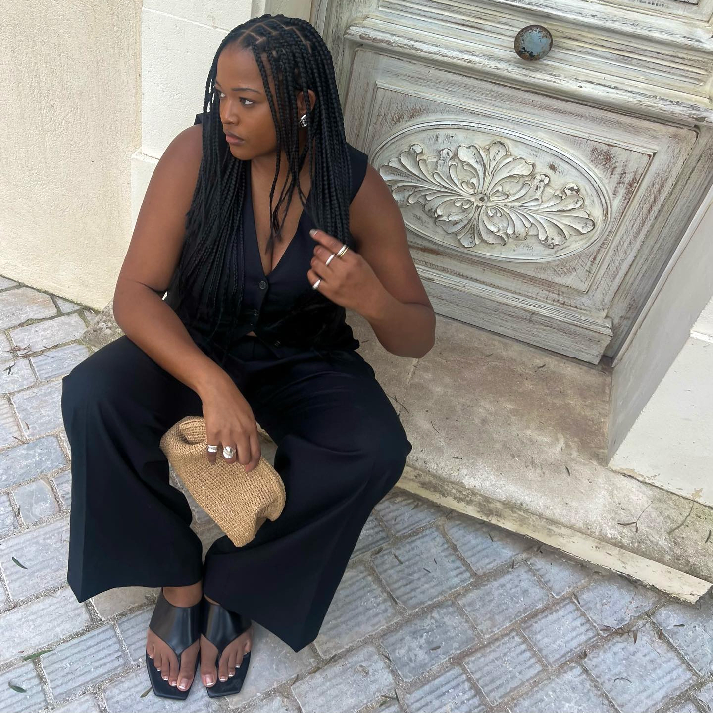 Imani Randolph sitting outside a beautiful European doorway wearing a black matching vest and trouser set, black flip-flop sandals, silver jewelry, and holding a raffia clutch.