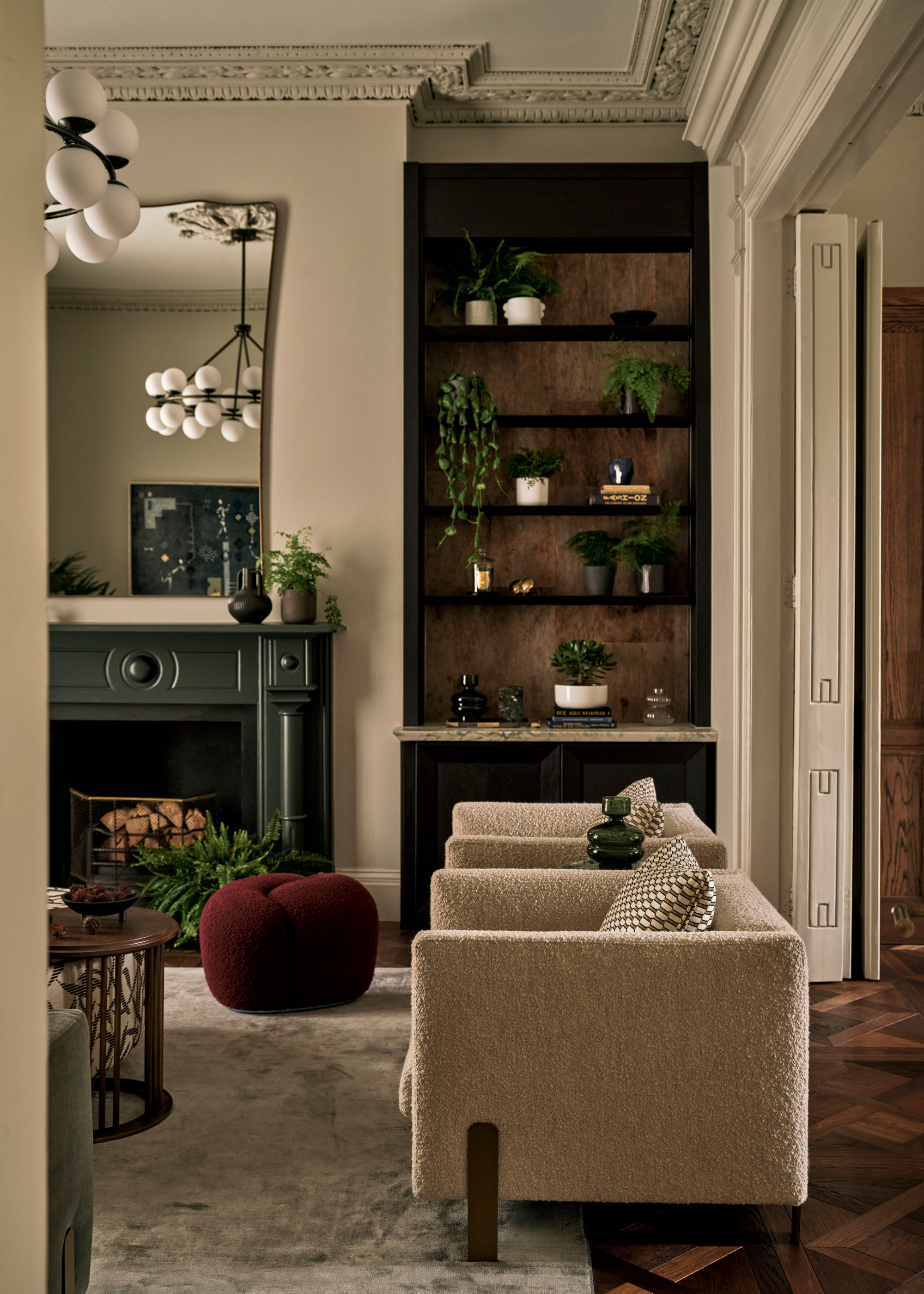 A white living room with a tall shelf of potted plants by a pair textured beige armchairs with patterned cushions by a maroon pouffe, a dark green fireplace below a tall mirror, and beside a coffee table