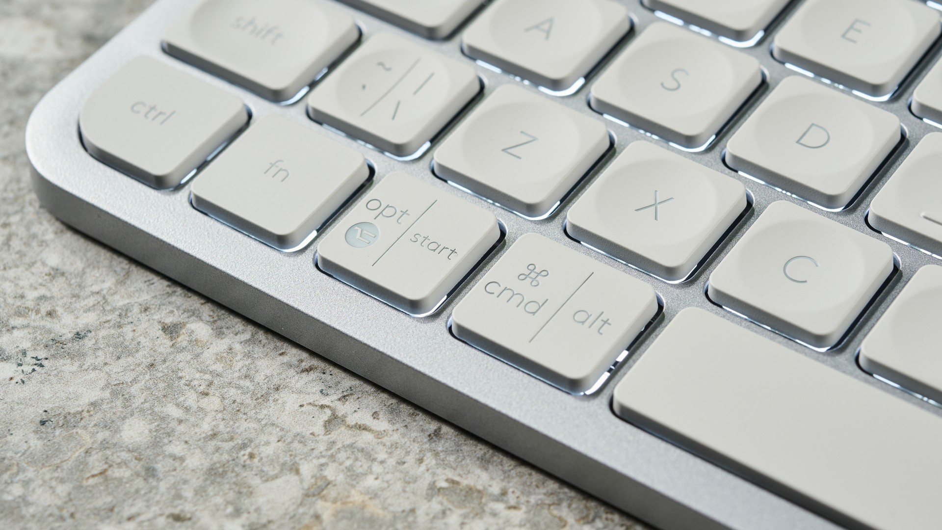 A photograph of the Logitech MX Keys Mini in light gray, positioned on a stone slab with a blue wall in the background.
