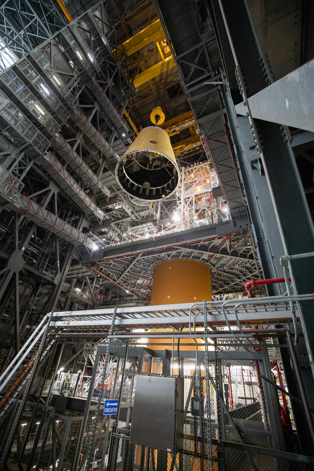 a large section of a rocket is hoisted in a factory above a tall orange rocket