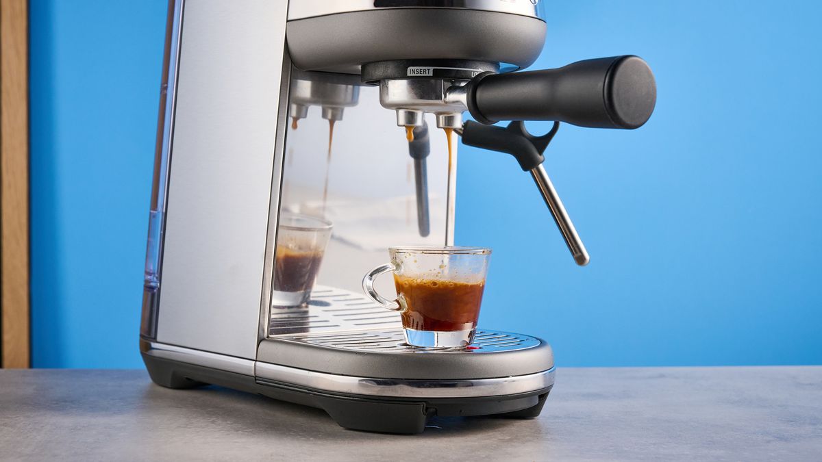 the breville bambino / sage bambino in silver photographed against a blue background with a wooden feature wall, showing the steam wand, group head, water tank, button controls, and espresso