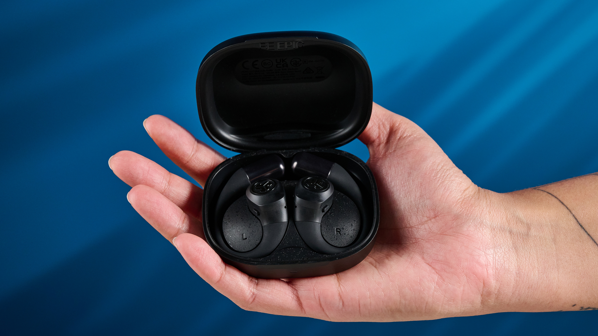 Jlab epic open sport open-earbuds in a black colorway photographed on a blue background.