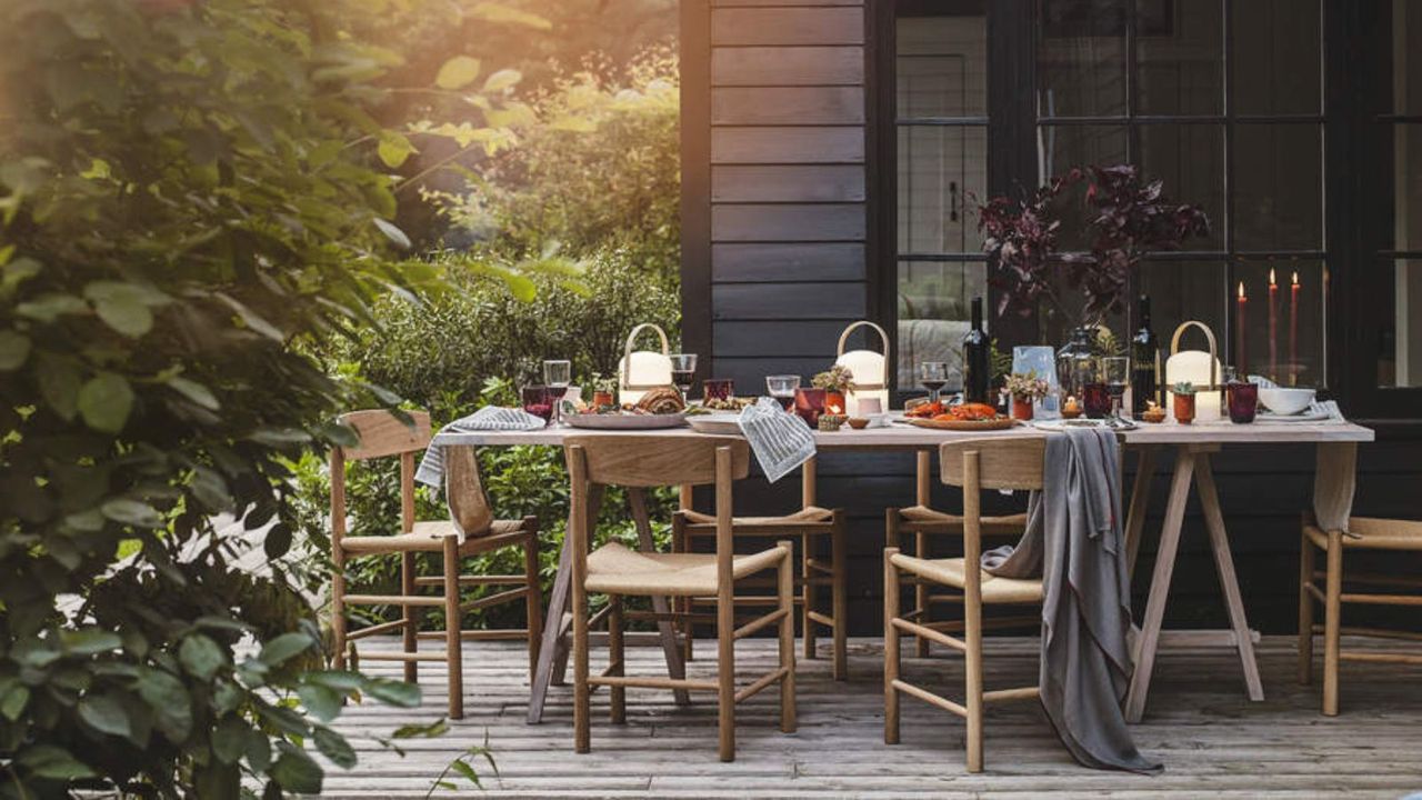 Outdoor dining area with wooden furniture decorated for a fall gathering with gray throws over the chairs and portable lamps along the table