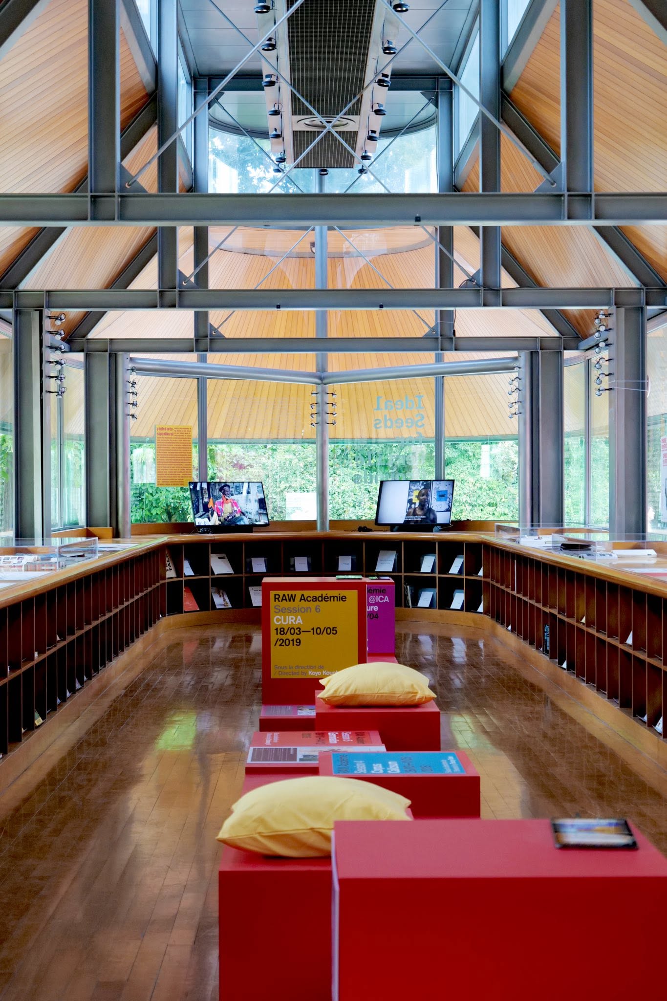 The wooden-clad interiors of a glassy gallery pavillion features an upside-down U structure with made-to-measure wooden shelving and floors, red cubical seats, and audiovisual installations.