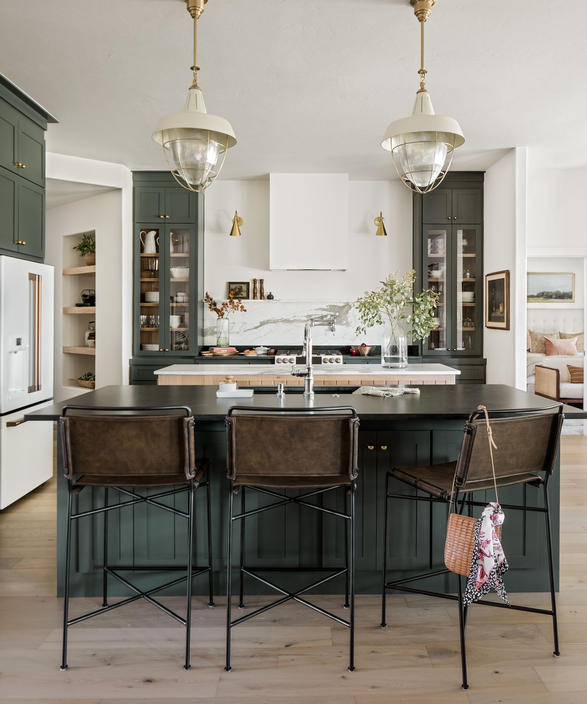 Kitchen with Pewter Green by Sherwin-Williams painted cabinets and island, marble backsplash, wooden floor and white walls, with two cream and gold pendants hanging from the ceiling.