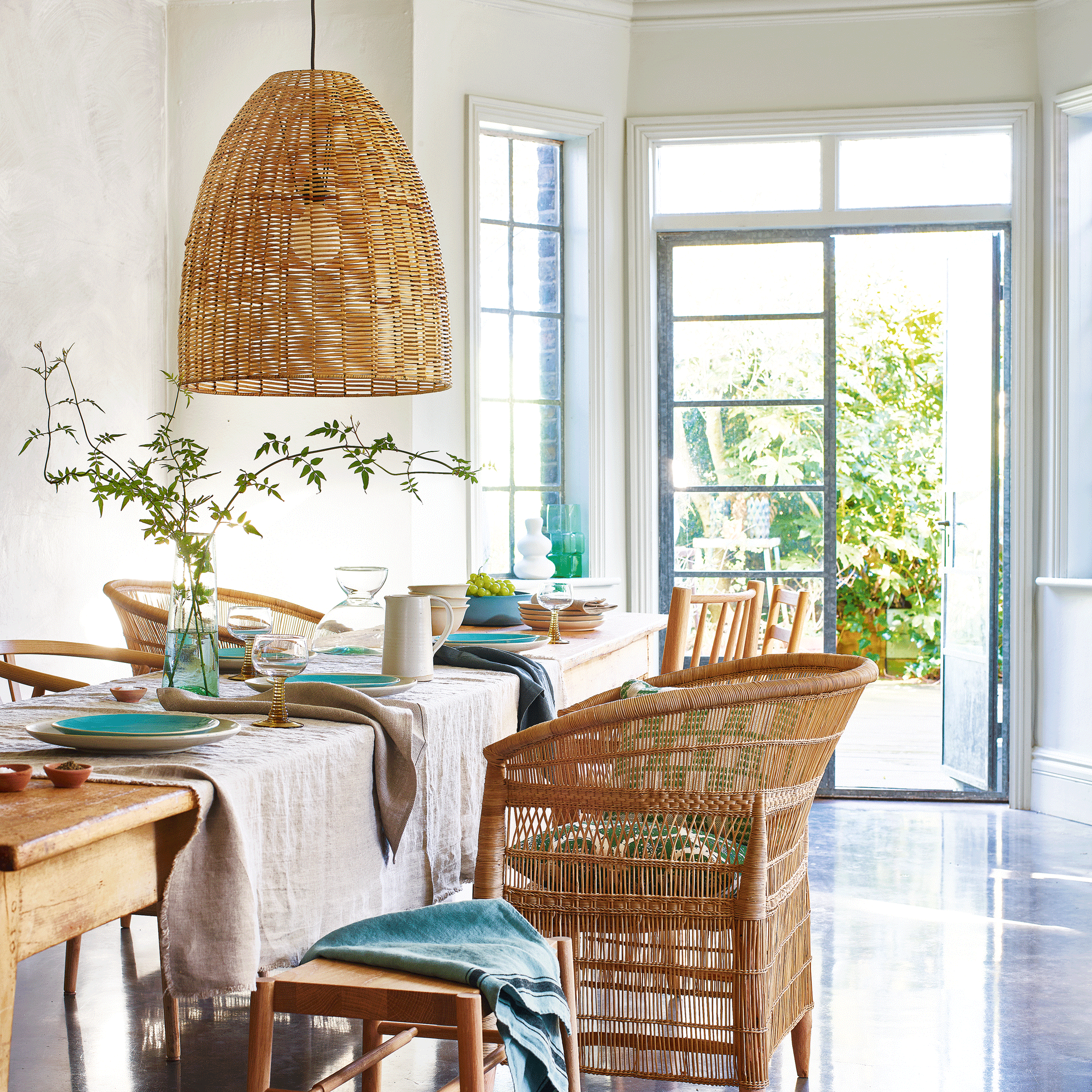 a dining room with table set for a meal and rattan chairs and a matching oversized rattan pendant overhead with open patio doors leading out into a garden